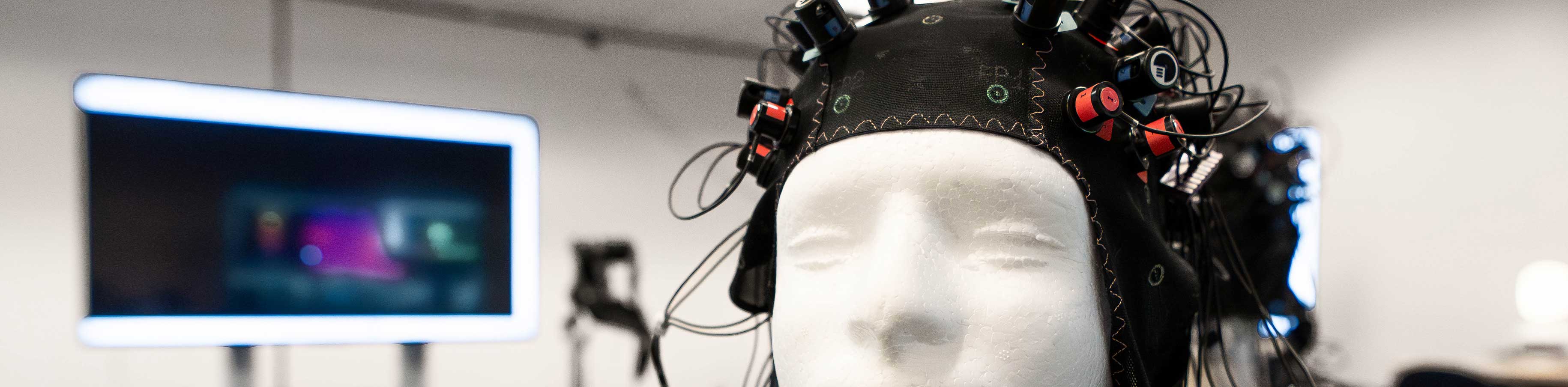 A mannequin head wearing a black EEG cap with wires is in a lab setting. A blurred screen in the background suggests a focus on technology and research.