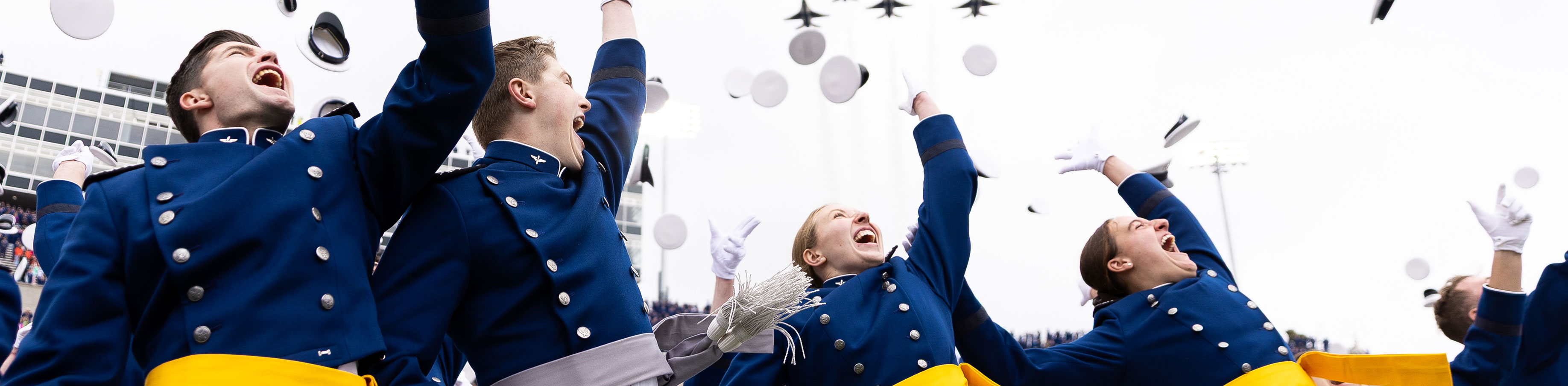 U.S. Air Force Academy graduates in blue uniforms joyfully throw caps in the air, celebrating outdoors. Jets fly overhead, adding a sense of excitement and achievement.
