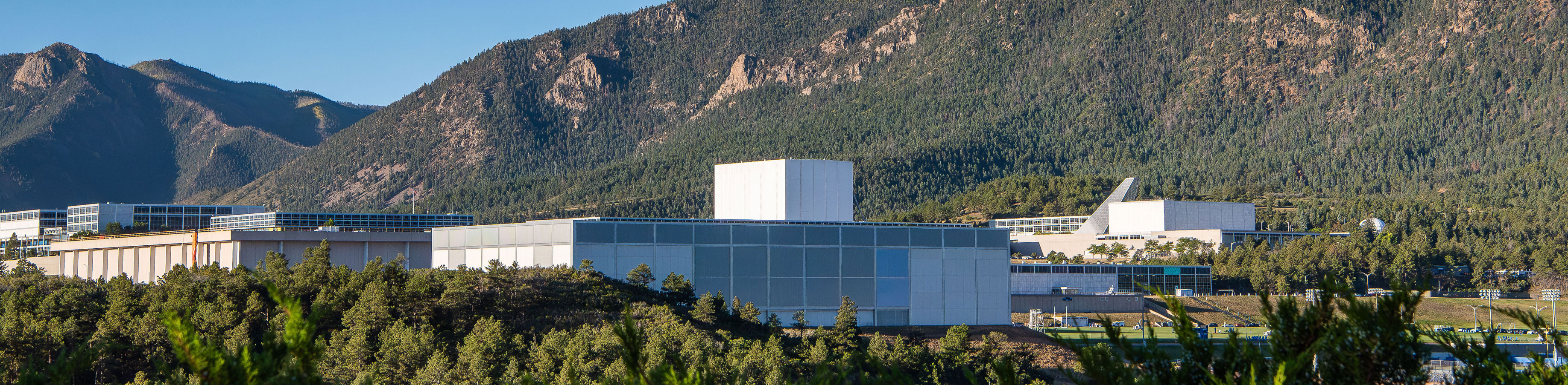 A view of the Holaday Athletic Center at the U.S. Air Force Academy with clean lines sit against a backdrop of forested mountains under a clear blue sky, conveying tranquility and harmony.