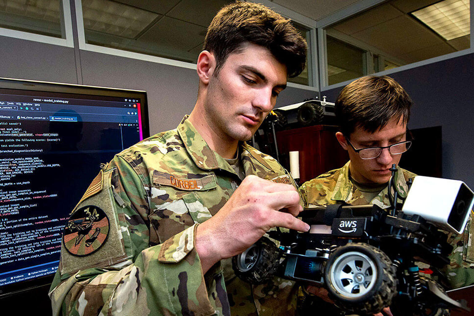 Two cadets in camouflage uniforms work on a small robotic vehicle indoors, with a computer displaying code in the background.