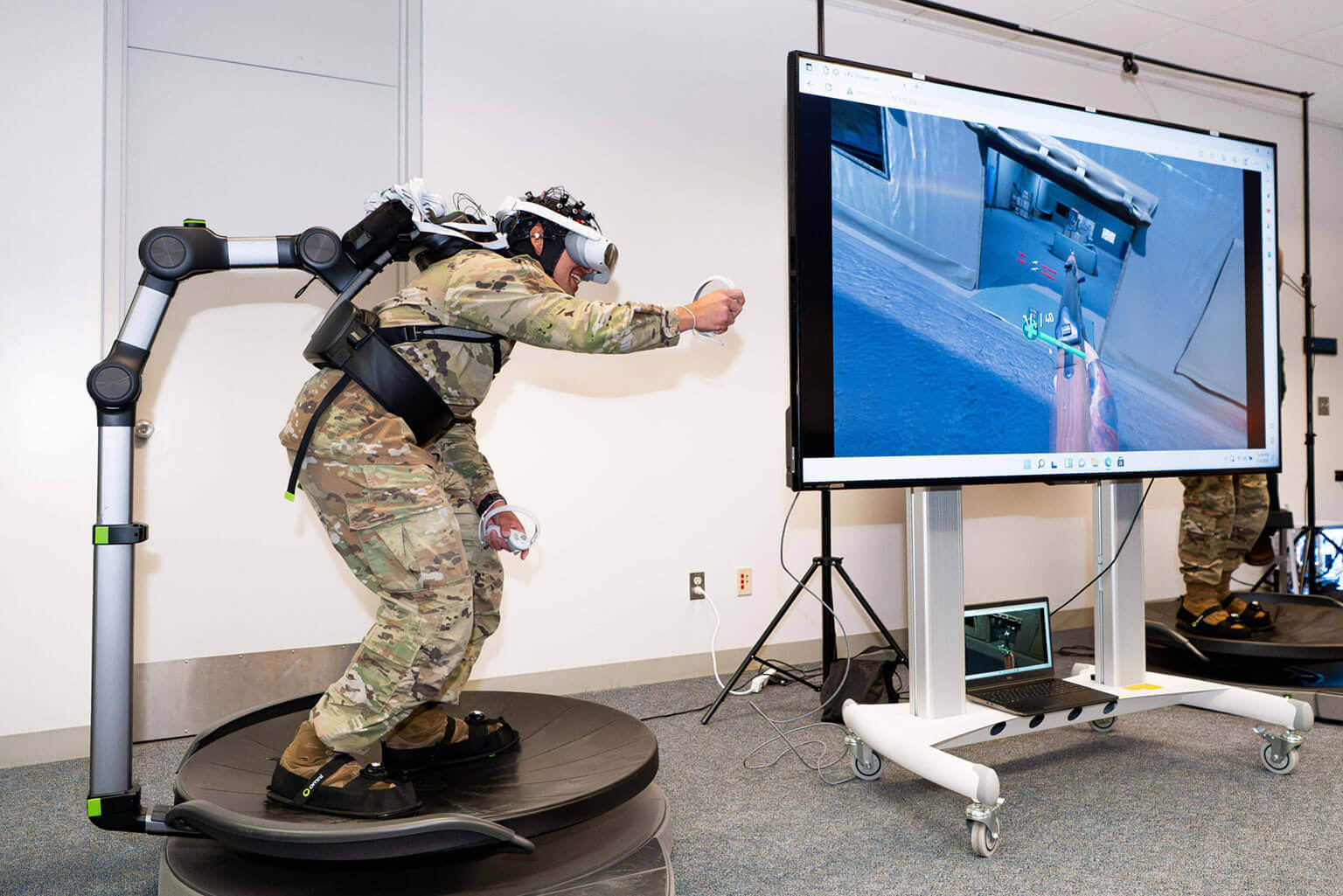 A cadet uses a VR headset and handheld controllers, standing on a motion platform, with a large screen displaying a virtual environment nearby.