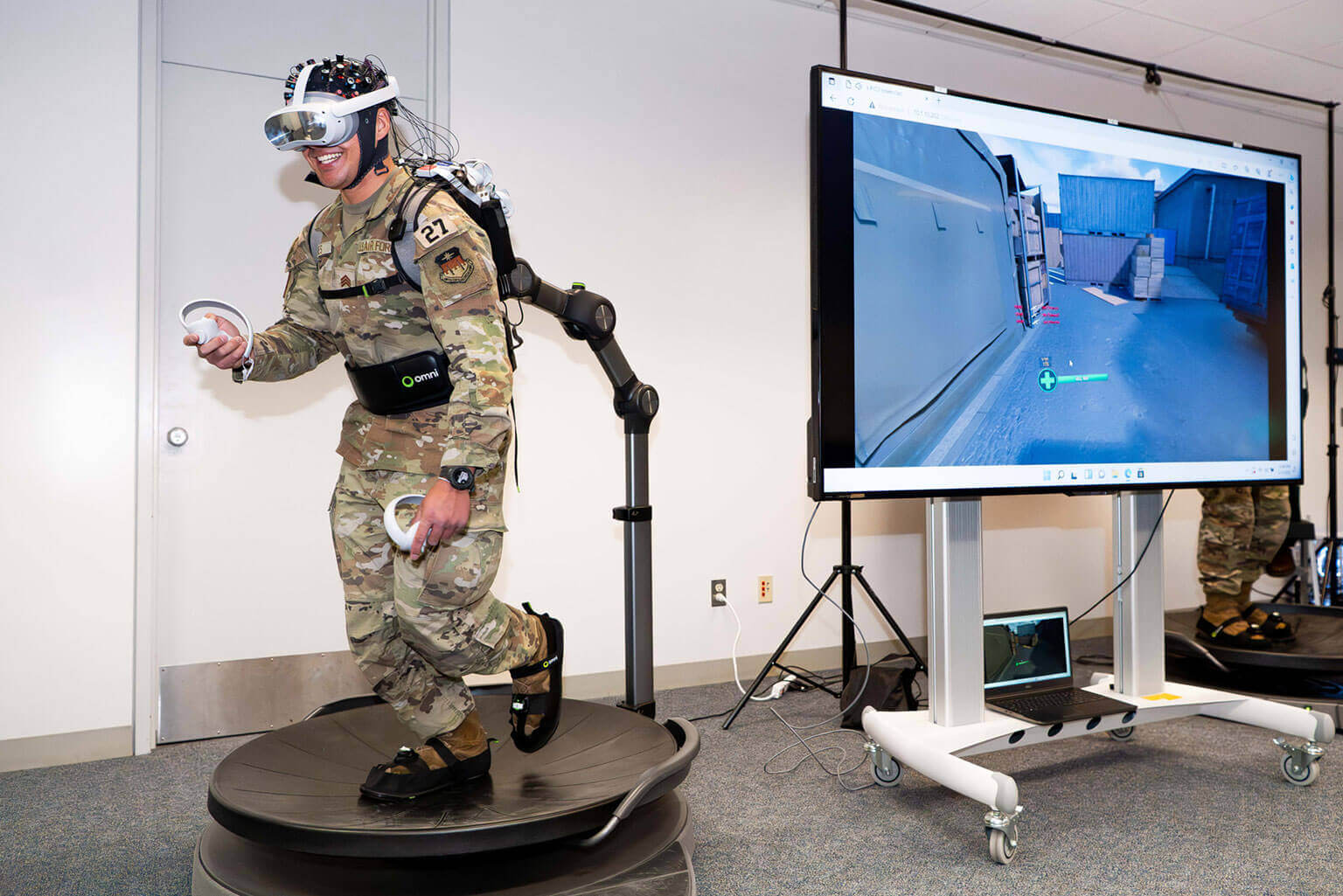 A cadet wearing a VR headset and uniform operates a virtual reality treadmill. A large screen displays a training simulation scene beside him.