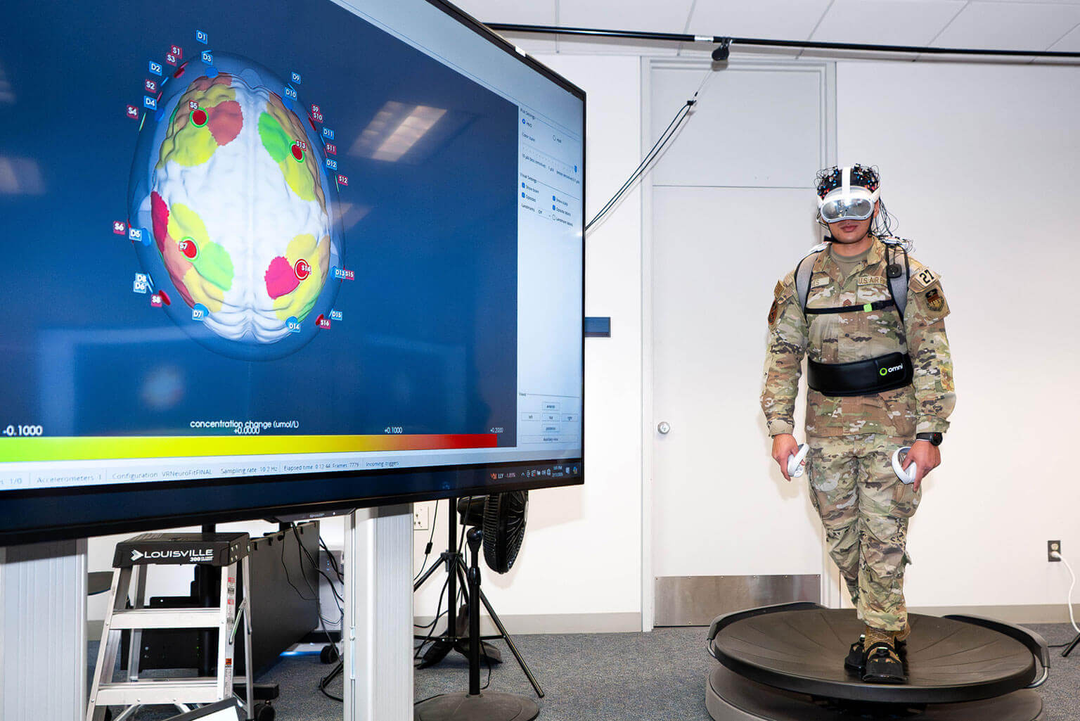 A cadet in camouflage and VR gear stands on a rotating platform. Nearby, a large screen displays a colorful, detailed brain scan. The setting is high-tech and experimental.