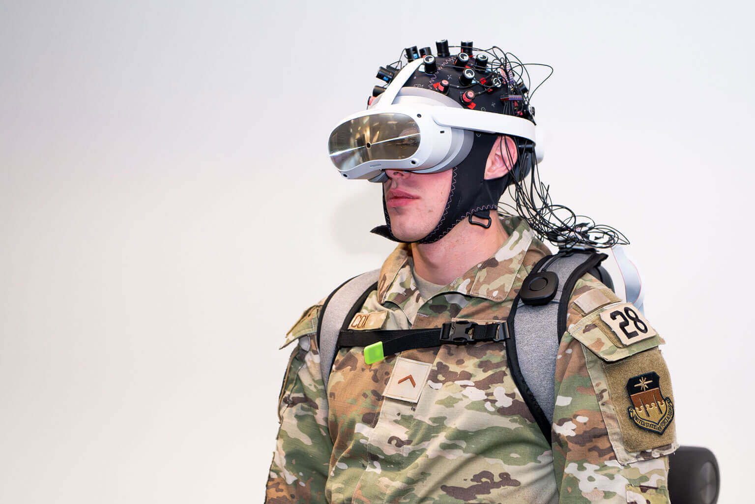 A cadet in camouflage military uniform wears a high-tech virtual reality headset with an attached neural sensor array, conveying a futuristic, focused look.