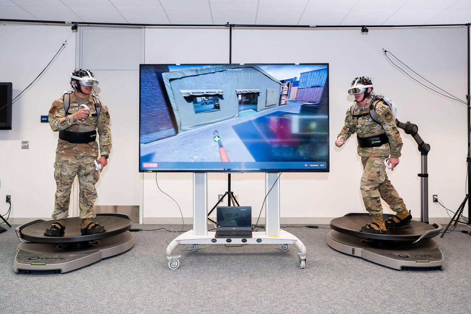 Two cadets in uniform use VR equipment and treadmills, engaging with a combat simulation displayed on a large screen. The atmosphere is focused.
