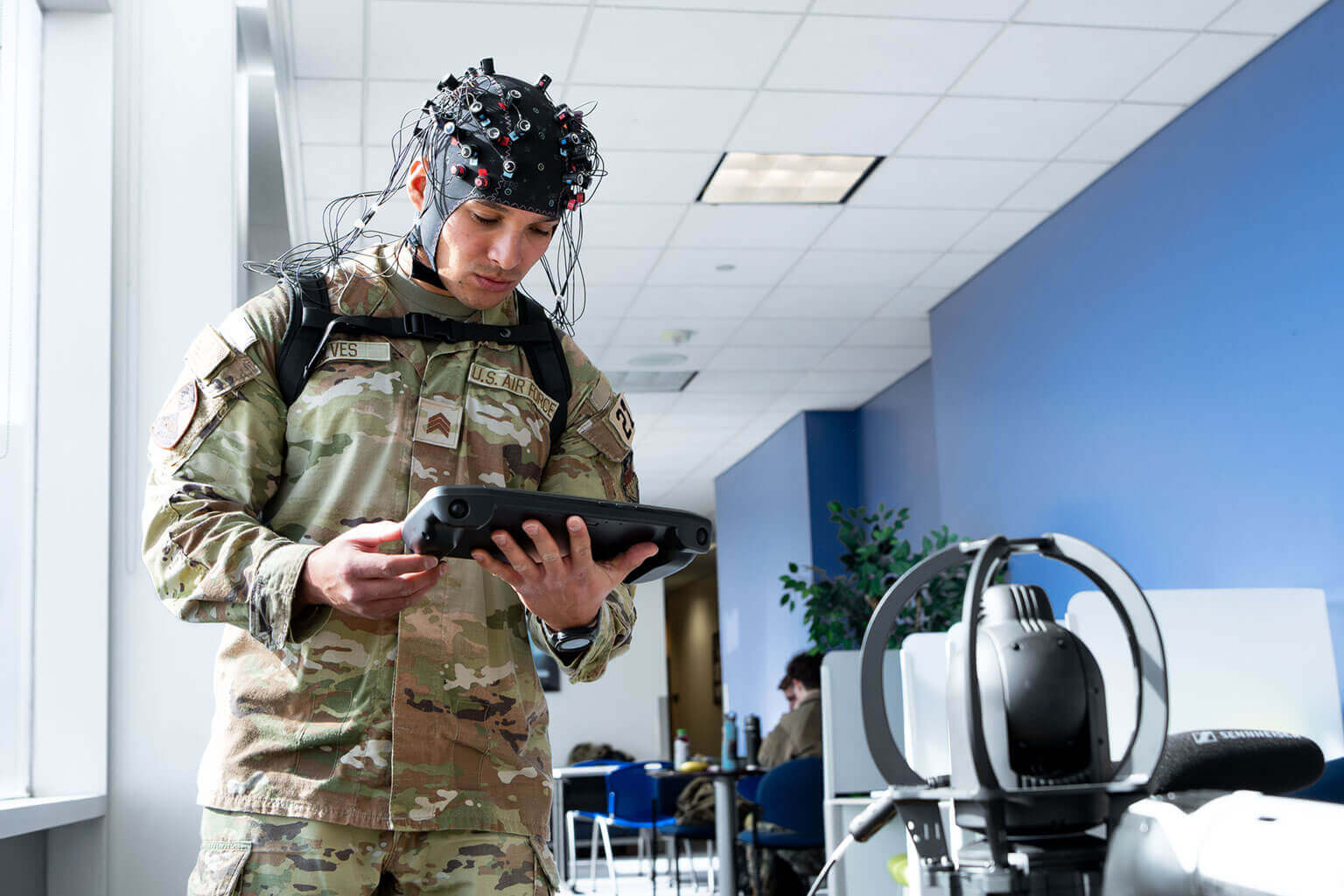 A cadet in military uniform wearing an EEG cap operates a tablet in a bright room, suggesting a focus on technology and research. Neutral tone.