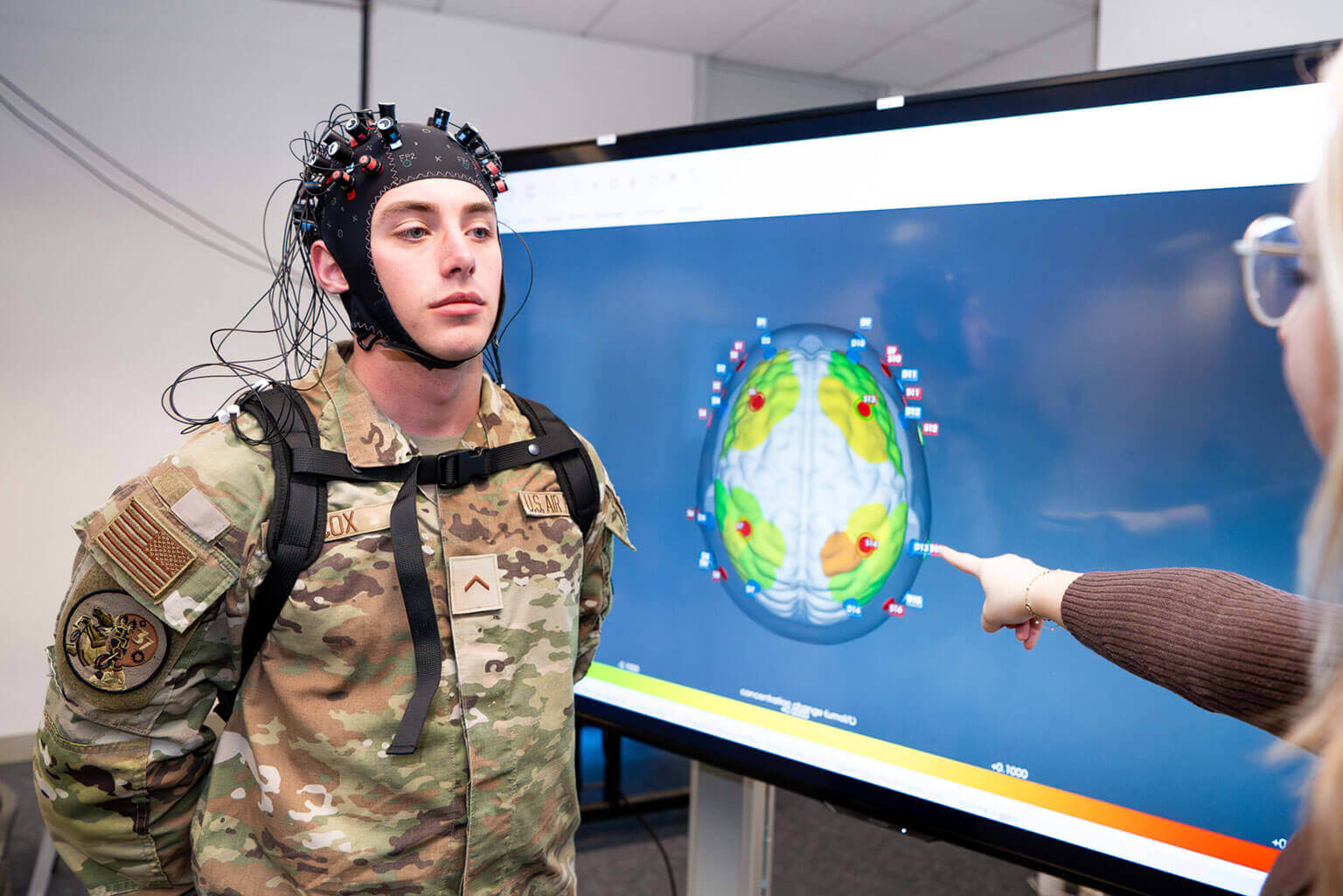 A cadet wearing a brain-monitoring cap stands beside a screen showing a colorful brain scan. A person points to the scan, indicating analysis.