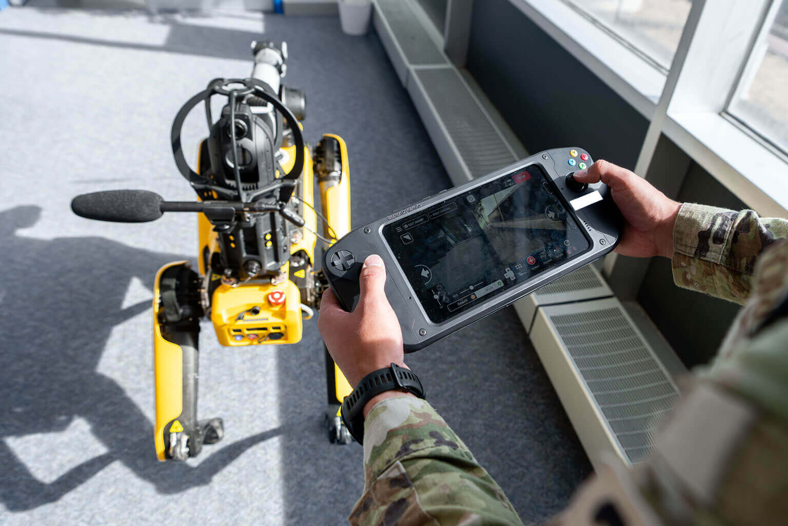 Cadet in camouflage remotely operates a yellow robotic dog using a handheld controller, indoors near large windows. The scene feels high-tech and controlled.