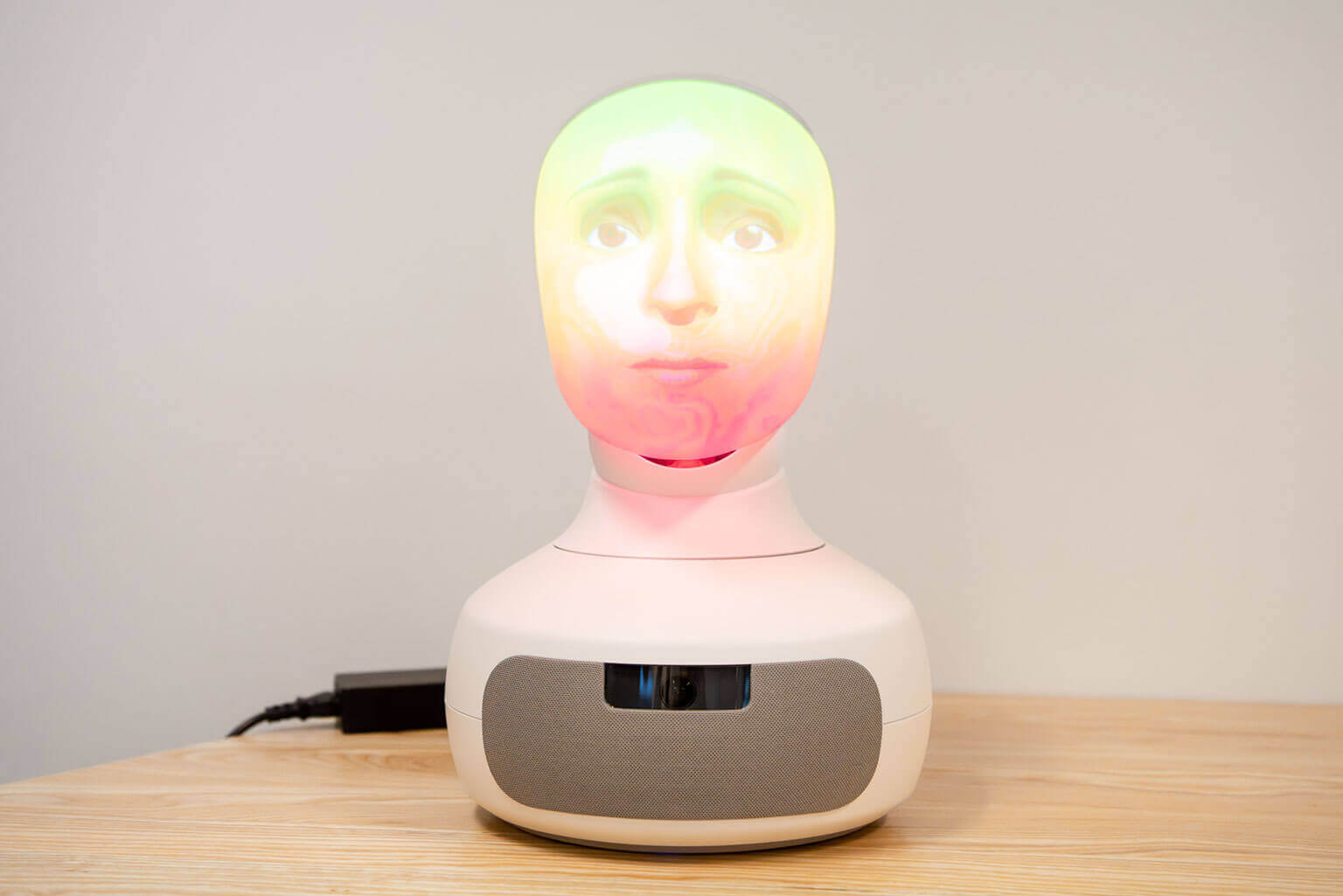 A futuristic robot with a glowing, colorful face displaying a serene expression is placed on a wooden table. The setting is minimalistic and modern.