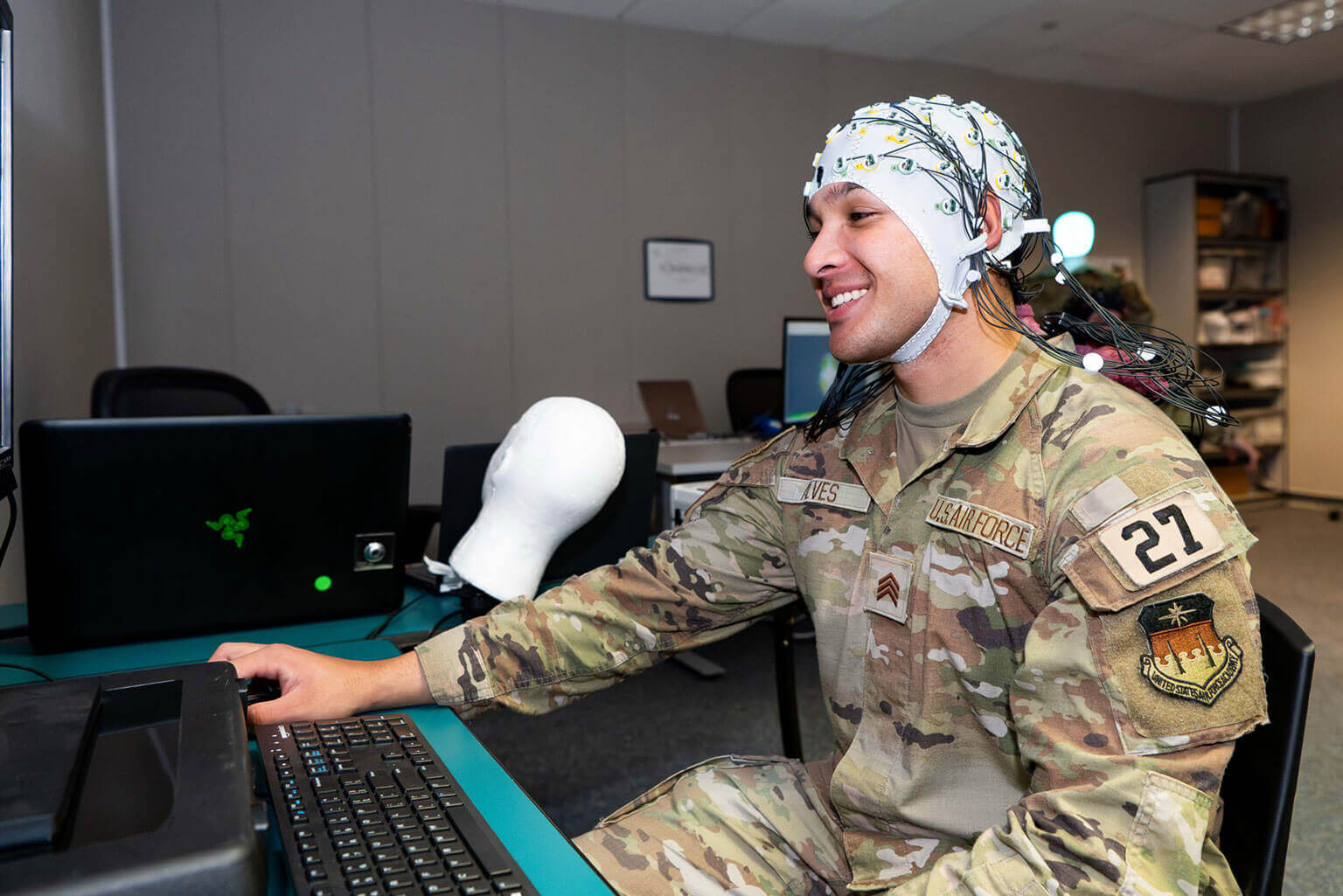 A cadet in military uniform smiles while using a computer. They're wearing an EEG cap connected to monitoring wires in a lab setting, indicating research or testing.