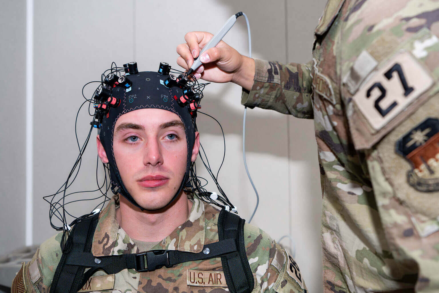 US Air Force Academy cadet in camouflage undergoes a brain scan, wearing a cap with electrodes. Another person adjusts equipment. Serious, focused atmosphere.