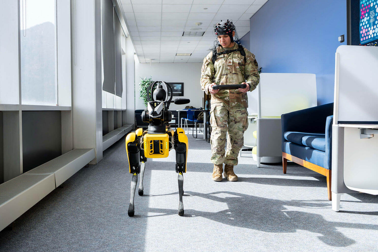 A cadet in military uniform operates a robotic dog using a remote control in a brightly lit room, suggesting advanced technology and innovation.