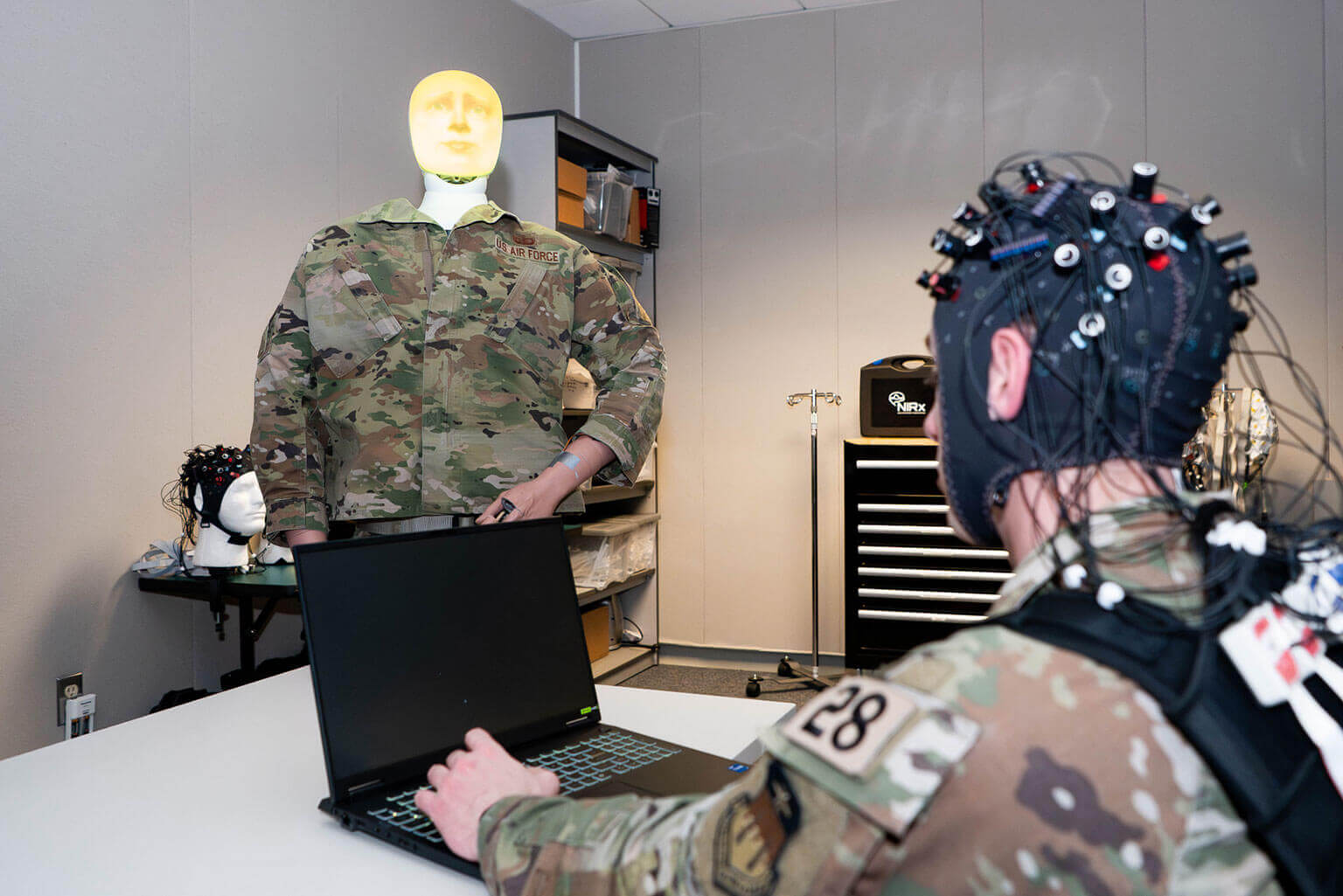 A cadet wearing brain-monitoring equipment sits at a desk with a laptop. A mannequin in camouflage stands nearby. The setting is a tech-filled lab.