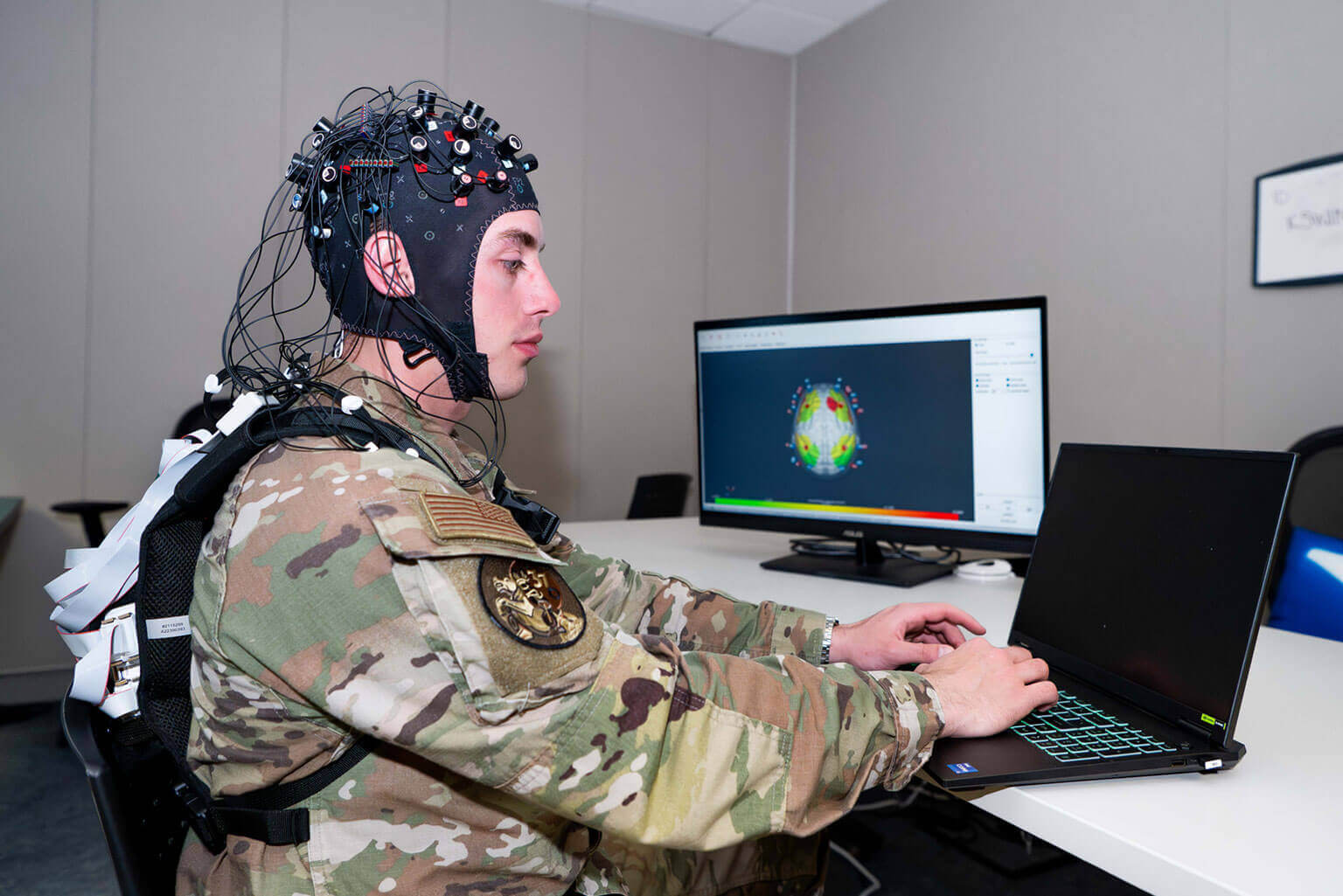 A cadet wearing a brain-monitoring EEG cap works on a laptop. A screen in the background displays brain activity data. The setting is a tech lab.