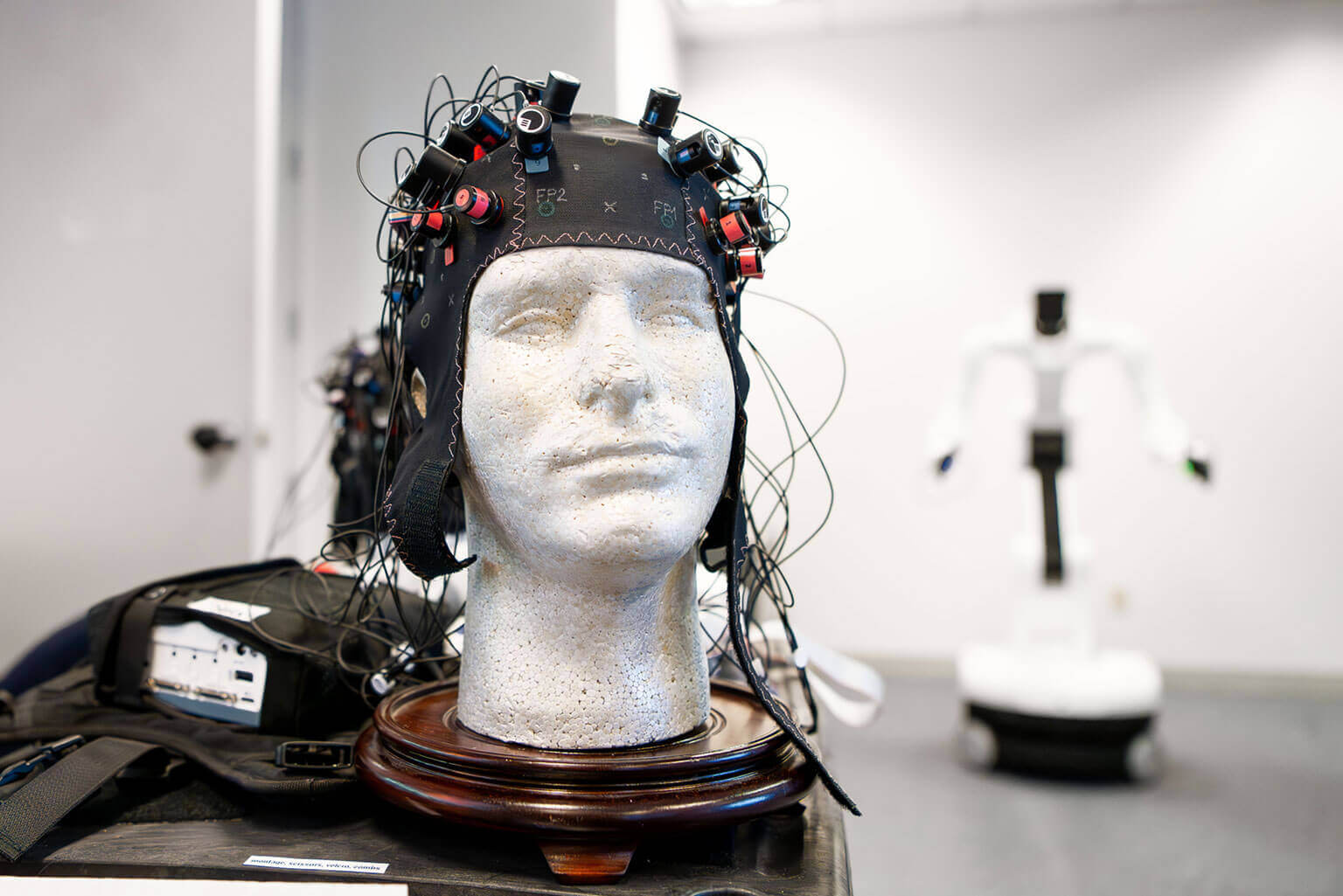 A mannequin head is fitted with a high-tech, sensor-laden cap, focusing on brain research. A humanoid robot is blurred in the background of a lab setting.