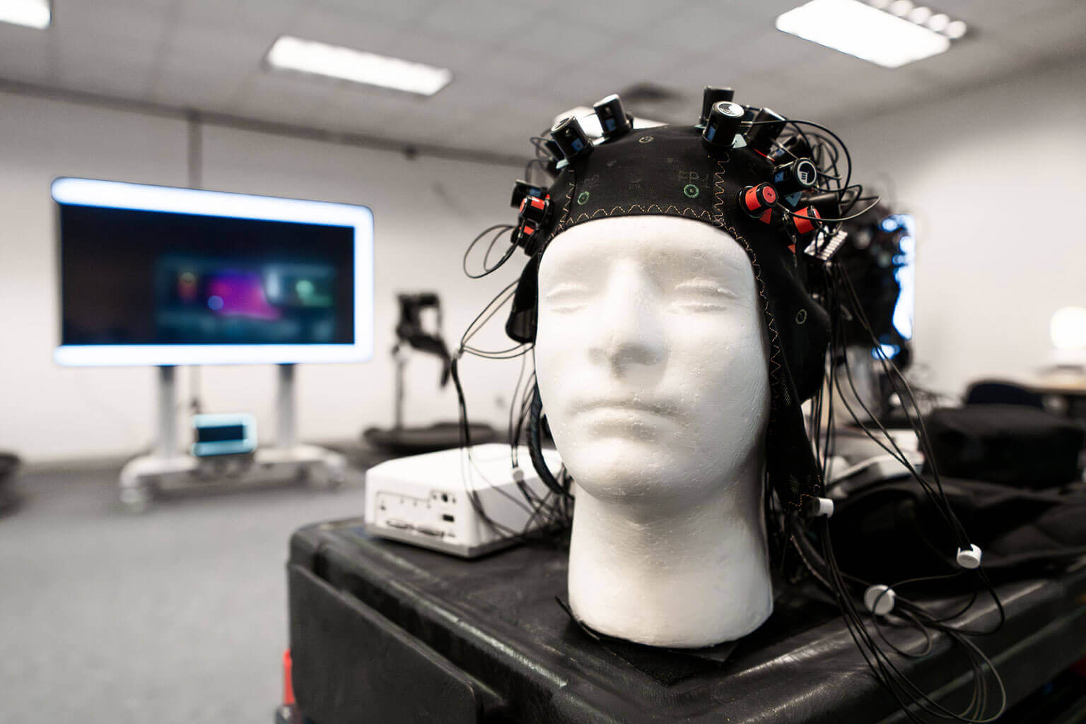 A mannequin head wearing an EEG cap with attached wires sits in the foreground of a tech lab. A large digital screen is blurred in the background.