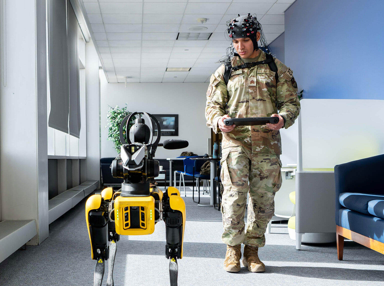 A cadet in uniform walks a robotic dog down a well-lit hallway, showcasing a moment of companionship and duty.