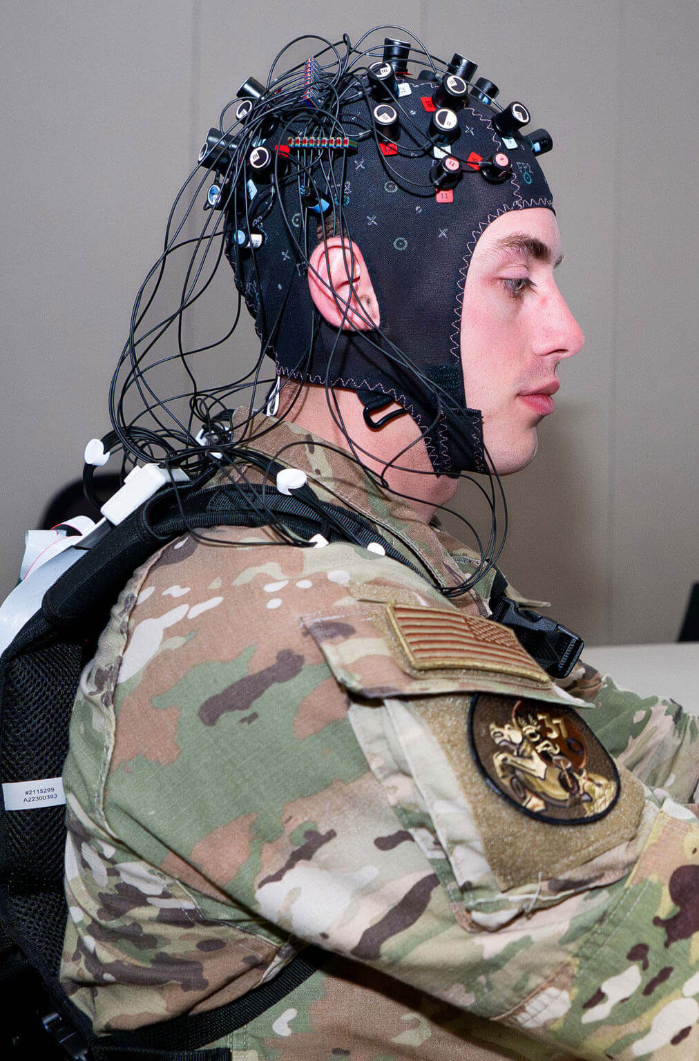 A cadet in camouflage uniform wearing a high-tech cap with numerous wires, illustrating advanced brain-interface technology. Focused expression, modern setting.